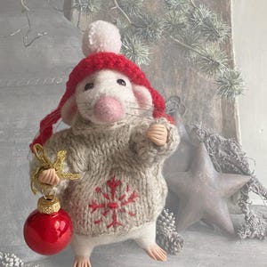 May include: A needle-felted white mouse figurine wearing a red knit hat with a pom-pom and a beige knit sweater featuring a red snowflake pattern. The mouse holds a small gold-trimmed red Christmas ornament in one hand and has pink ears.