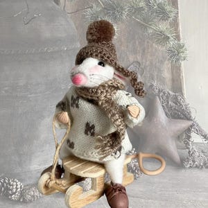 May include: A white felt mouse figurine wearing a brown crocheted hat with a pom-pom, a matching brown crocheted scarf, and a cream-colored sweater with brown patterns. The mouse is positioned on a small wooden sled with a pull rope.