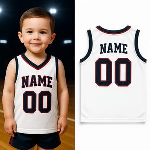 Puede incluir: Una camiseta de baloncesto blanca con ribetes azul marino y rojos. La camiseta muestra el nombre "NAME" y el n&uacute;mero "00" en azul marino con detalles rojos. La imagen muestra la camiseta en un ni&ntilde;o peque&ntilde;o y una vista separada de la camiseta.