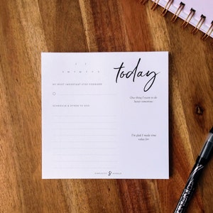 May include: A white notepad with the word "today" in cursive script. The notepad includes sections for a daily schedule, a most important step forward, and a space to write what you are glad you made time for. A black pen is in the foreground.