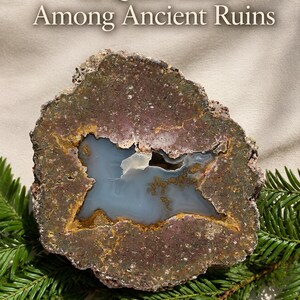 May include: A geode with a rough, dark brown and purple outer crust surrounding a smooth, light blue crystalline center. The text "A Quiet Lake Among Ancient Ruins" is printed above the stone, which rests on green pine needles.