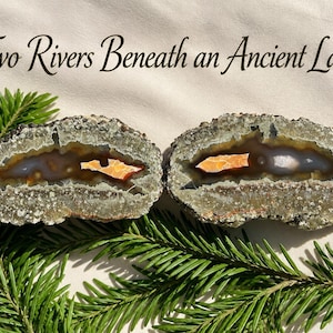 May include: Two geode halves with rough, grey, crystalline outer edges and smooth, brown, translucent interiors featuring bright orange, jagged, central inclusions. The text "Two Rivers Beneath an Ancient Land" is printed above the stones.