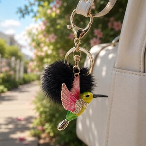 May include: A gold-coloured hummingbird charm with pink and green enamel, embellished with rhinestones, hangs from a keychain with a black pom-pom. The charm is attached to a white handbag with a gold clasp.
