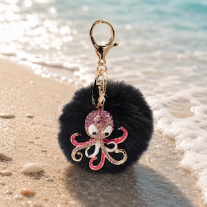 May include: A black fluffy keychain with a gold-coloured clasp. The keychain features a bejewelled octopus charm with pink and white accents. The octopus has a gold outline against a sandy beach background.