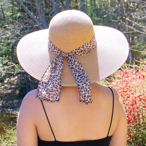 May include: A woman wearing a wide-brimmed straw hat with a leopard print ribbon tied in a bow at the back. She is wearing a black sundress and is standing in a grassy area.