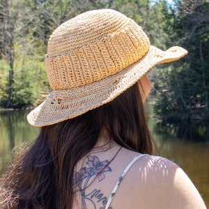 May include: A woman with long brown hair wearing a straw hat with a wide brim. She has a tattoo on her back with a floral design and the text "Never" and "My blood" with the date "1-14-19".