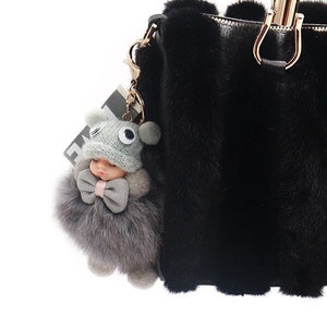 Op de afbeelding: Zwarte handtas van imitatiebont met gouden hardware en een pluche grijze speelgoed sleutelhanger. De tas heeft een ritssluiting bovenaan en twee bovenste handvatten.