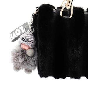 Puede incluir: Un bolso de mano de piel sintética negra con herrajes dorados y un llavero gris esponjoso con la palabra "LOVE" en él.