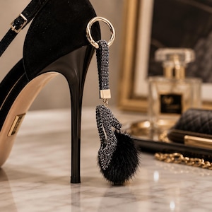 May include: A black high-heeled shoe with a matching charm. The charm features a miniature high heel adorned with rhinestones and a fluffy black pom-pom. A gold ring attaches the charm to the shoe.