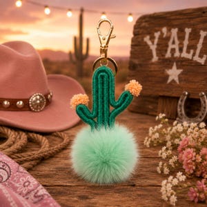 May include: A green cactus keyring with a gold clasp and orange pom-pom details. The cactus sits on a fluffy, light green pom-pom. The background includes a pink cowboy hat, a wooden sign that says "Y'all", and a horseshoe.