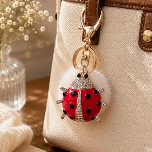 May include: A red ladybug keyring with black spots and rhinestone detailing hangs from a gold clasp attached to a cream handbag. The ladybug has a fluffy white pom-pom base.