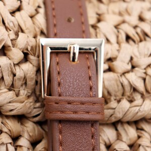 May include: Close-up of a brown leather strap with a gold-colored metal buckle. The strap is stitched with light brown thread and is set against a woven straw background. The buckle is square and has a single bar.
