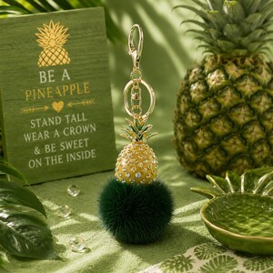 May include: A gold pineapple keyring with a green fluffy base. The pineapple is yellow with crystal accents and a green top. A wooden sign with the text "BE A PINEAPPLE" is in the background.