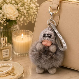 May include: A gray fur pom-pom keychain with a sleeping baby doll wearing a gray knit hat with "HDSP" on it. The keychain has a silver clasp and a gray strap that says "LOVE".