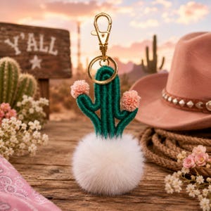 Peut inclure: Un porte-clés en forme de cactus vert avec des pompons roses et une base duveteuse blanche. Le porte-clés a un fermoir et un anneau dorés. L'arrière-plan comprend une enseigne en bois avec l'inscription "Y'ALL", un chapeau de cow-boy rose et un paysage désertique.