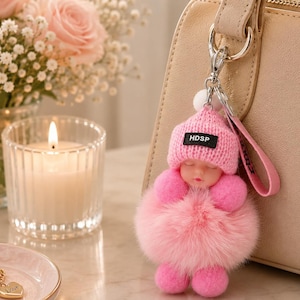 May include: A pink baby doll keychain with a knitted hat that reads "HDSP" is attached to a beige purse. The doll has a fluffy pink body and is next to a lit candle, a perfume bottle, and a bouquet of pink roses.