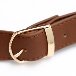 May include: Close-up of a brown belt with a gold-colored buckle. The belt is made of a smooth, leather-like material with visible stitching. The gold buckle has a classic design.