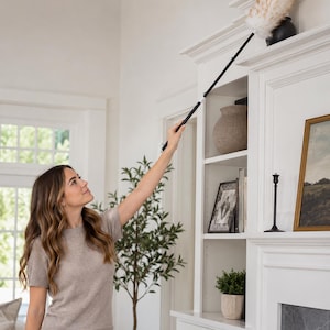 May include: A woman using a long-handled duster with a fluffy, light-colored head to clean a shelf. The duster is black and extends to reach high places. The woman is wearing a light brown short-sleeved shirt and blue jeans.