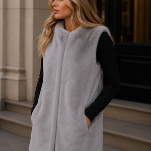 May include: A light gray faux fur vest with a rounded collar and two front pockets. The vest is open in the front and appears to be knee-length. The model is wearing a black long-sleeve shirt underneath.