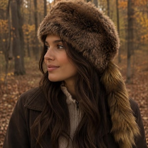 Peut inclure: Un chapeau en fausse fourrure marron avec une longue queue, porté par une personne dans une veste marron. Le chapeau est un accessoire d'hiver chaud. L'arrière-plan est une scène de forêt floue avec des couleurs d'automne.