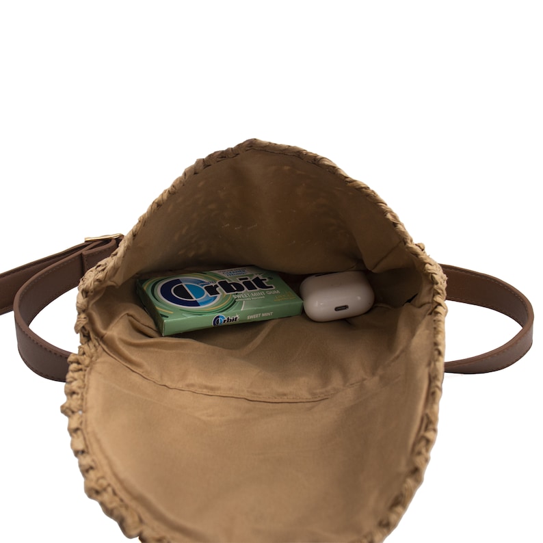 May include: Open view of a woven straw shoulder bag with a brown leather strap. Inside the bag are a pack of Orbit Sweet Mint gum and a pair of white wireless earbuds. The bag is tan and has a brown leather strap.