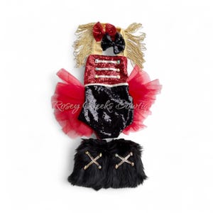 May include: A baby outfit with a red sequined top, black sequined bottom, and a red tulle skirt. The outfit includes a gold headband with a red bow and black bow, and black faux fur leg warmers with gold cross accents.