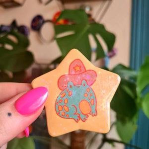 May include: A yellow star-shaped pin with a green frog wearing a red cowboy hat with a yellow star. The frog has orange spots and is surrounded by a glittery background.