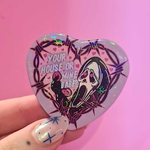 May include: A heart-shaped button with a holographic finish features a black and white illustration of a screaming ghost with a barbed wire border. The text "YOUR HOUSE OR MINE BABE?" is printed on the button.