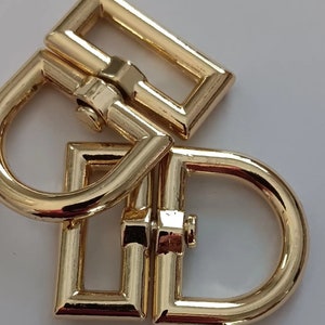 May include: Gold metal buckle with a D-ring. The buckle has a rectangular shape with a smooth, polished finish.
