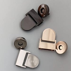 May include: Three magnetic closures for bags or purses. The closures are in three different finishes: black, gold, and silver. Each closure has a round magnetic button and a rectangular metal piece with a curved top.