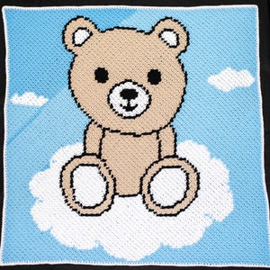 May include: A crocheted baby blanket with a brown teddy bear sitting on a white cloud against a blue sky background.