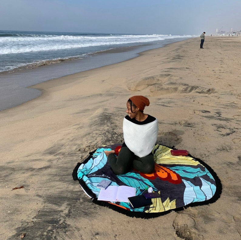 May include: A person sitting on a beach with a colorful round blanket, wearing a white sweater and a brown beanie. There are notebooks and a phone on the blanket.
