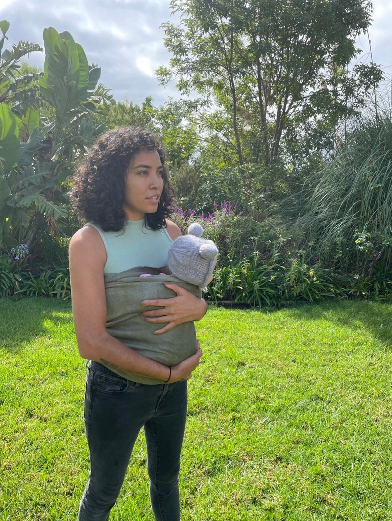 May include: A woman wearing a green wrap carrier with a baby inside. The baby is wearing a gray hat with bear ears. The woman is standing in a grassy area with trees in the background.
