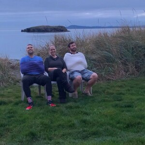 May include: Three people are sitting on white chairs on a grassy hill overlooking a body of water. The people are wrapped in blankets and are looking out at the water.