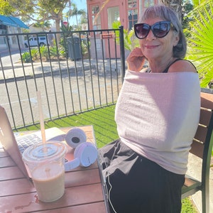 May include: A woman wearing sunglasses and a light pink shirt sits at a table with a laptop, a cup of coffee, and white headphones. The table is made of wood and has a brown finish. The woman is sitting on a chair with a black seat and a brown back.