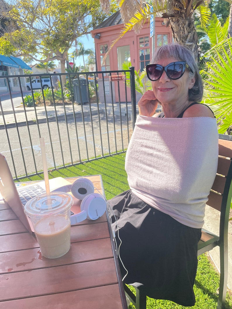 May include: A woman wearing sunglasses and a light pink shirt sits at a table with a laptop, a cup of coffee, and white headphones. The table is made of wood and has a brown finish. The woman is sitting on a chair with a black seat and a brown back.
