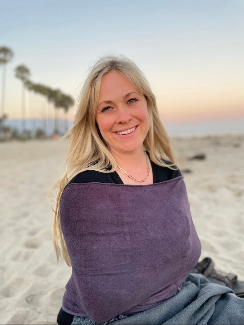 May include: A woman wearing a purple hooded towel on a beach. She is smiling and looking at the camera.
