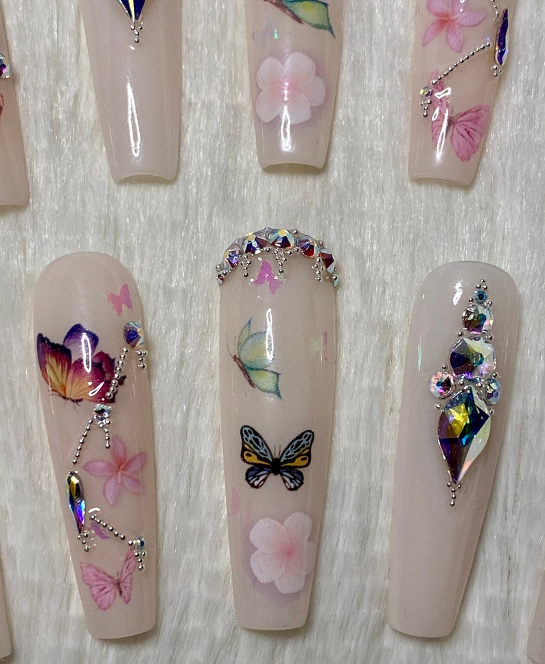 Pu&ograve; includere: Un set di unghie finte lunghe, rosa chiaro. Ogni unghia presenta disegni di farfalle e fiori, alcune decorate con strass iridescenti. Le unghie hanno una finitura lucida, creando un'estetica delicata e femminile.