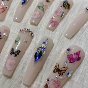 Pu&ograve; includere: Un set di unghie finte lunghe, rosa chiaro. Ogni unghia presenta decalcomanie di farfalle e fiori, alcune decorate con strass iridescenti. Le unghie hanno una finitura lucida e una forma rettangolare.