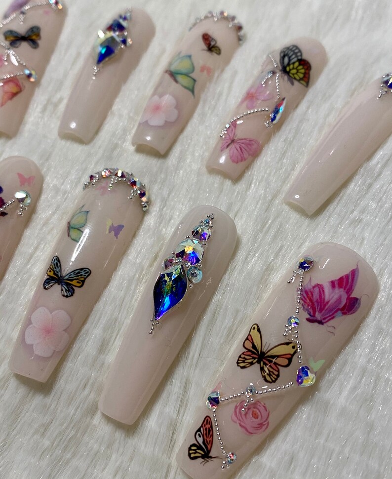 Pu&ograve; includere: Un set di unghie finte lunghe, rosa chiaro. Ogni unghia presenta decalcomanie di farfalle e fiori, alcune decorate con strass iridescenti. Le unghie hanno una finitura lucida e una forma rettangolare.