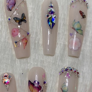 Pu&ograve; includere: Un set di unghie finte in una tonalit&agrave; rosa chiaro, decorate con decalcomanie a farfalla, strass e dettagli floreali. Alcune unghie presentano una cascata di strass, altre un'unica gemma.