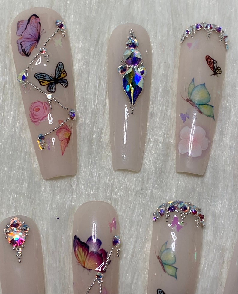 Pu&ograve; includere: Un set di unghie finte in una tonalit&agrave; rosa chiaro, decorate con decalcomanie a farfalla, strass e dettagli floreali. Alcune unghie presentano una cascata di strass, altre un'unica gemma.