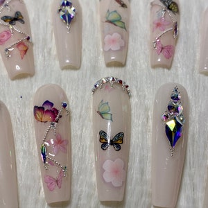 Pu&ograve; includere: Un set di unghie finte lunghe, rosa chiaro. Alcune unghie presentano decalcomanie di farfalle e fiori, mentre altre sono decorate con strass scintillanti. Le unghie sono disposte su una superficie bianca e soffice.