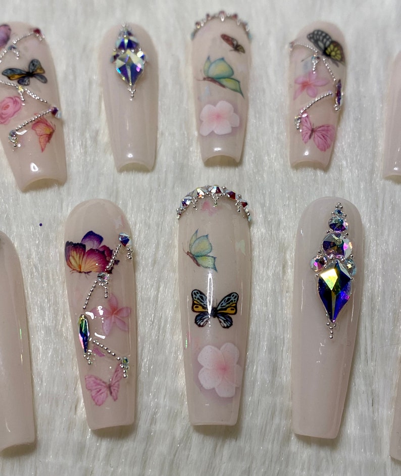 Pu&ograve; includere: Un set di unghie finte lunghe, rosa chiaro. Alcune unghie presentano decalcomanie di farfalle e fiori, mentre altre sono decorate con strass scintillanti. Le unghie sono disposte su una superficie bianca e soffice.