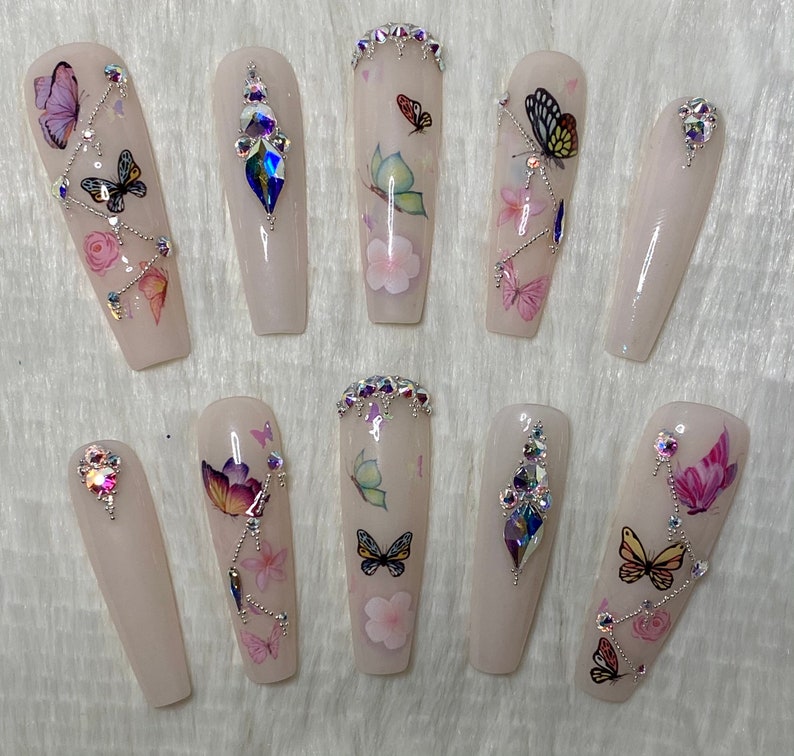 Pu&ograve; includere: Set di dieci unghie finte lunghe a forma di bara. Ogni unghia &egrave; rosa pallido con decalcomanie di farfalle e fiori. Alcune unghie presentano strass e gemme iridescenti. Le farfalle sono di vari colori.