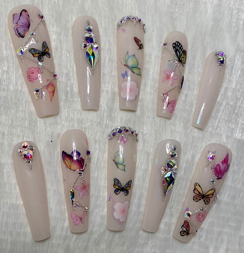 Pu&ograve; includere: Un set di dieci unghie finte lunghe a forma di bara. Ogni unghia &egrave; rosa pallido con decalcomanie di farfalle e fiori. Alcune unghie presentano strass iridescenti e dettagli a catena argentata. Le farfalle sono in vari colori.