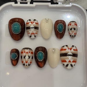 Puede incluir: Un juego de diez uñas artificiales en una caja de plástico blanco. Las uñas presentan varios diseños, incluyendo purpurina marrón con una piedra turquesa, patrones geométricos y uñas de color crema sólido con una pequeña gema.