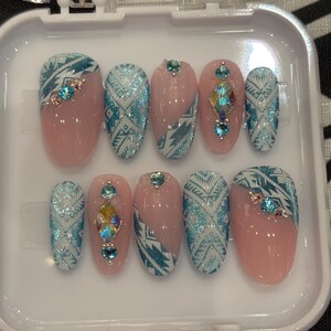 Puede incluir: Un juego de diez uñas artificiales en una caja de plástico transparente. Las uñas presentan una base rosa claro con patrones geométricos turquesa y blancos y gemas iridiscentes. Las uñas son de forma ovalada.