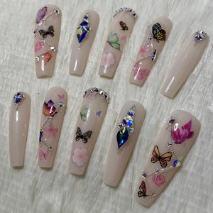 Pu&ograve; includere: Un set di dieci unghie finte lunghe, rosa chiaro. Ogni unghia presenta decalcomanie di farfalle e fiori, strass argentati e gemme iridescenti. Le unghie sono disposte su una superficie bianca e soffice.