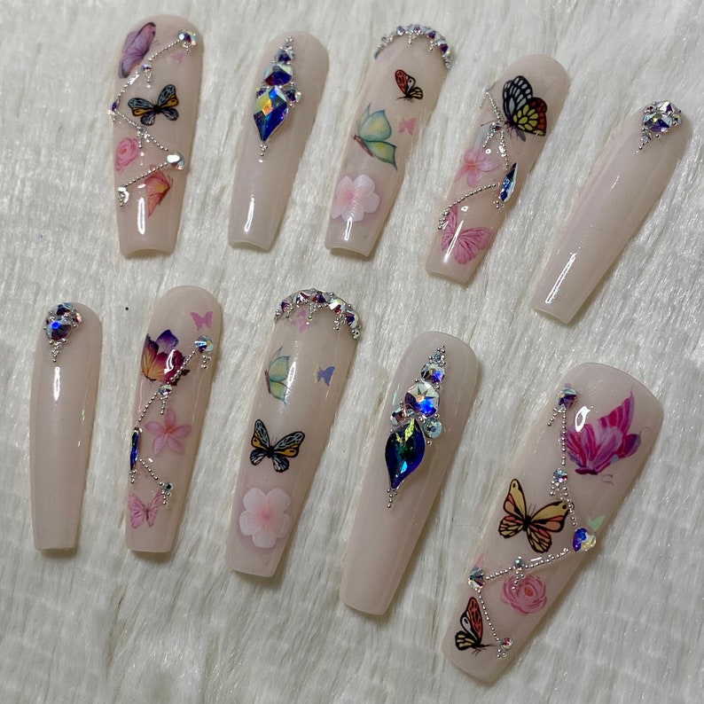 Pu&ograve; includere: Un set di dieci unghie finte lunghe, rosa chiaro. Ogni unghia presenta decalcomanie di farfalle e fiori, strass argentati e gemme iridescenti. Le unghie sono disposte su una superficie bianca e soffice.